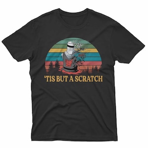 May include: Black t-shirt featuring a graphic of a knight in armor with a distressed effect, set against a sunset backdrop. The text "'TIS BUT A SCRATCH" is printed below the graphic. The design is a humorous reference to a classic movie scene.