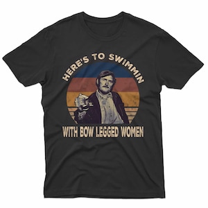 May include: Black t-shirt featuring a vintage-style graphic of a man holding a drink, set against a sunset-like background. The text reads "HERE'S TO SWIMMIN WITH BOW LEGGED WOMEN."