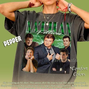 May include: Dark grey unisex t-shirt with the name "Nathan Fillion" in green, and images of the actor. The shirt has the word "Pepper" in a white box and "Comfort Colors" in the bottom right corner.