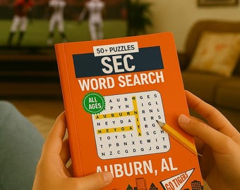 May include: An orange "SEC Word Search" puzzle book with the text "Auburn, AL" and "Go Tigers". The book is being held with a pencil, with a football game playing on a TV in the background.
