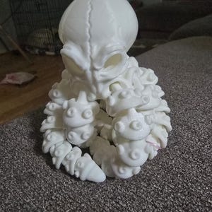 May include: A white, 3D-printed alien sculpture with a large head, prominent eyes, and detailed tentacles. The sculpture is made of a smooth, solid material and sits on a textured gray surface. The alien figure is a unique decorative item.