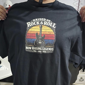 May include: Black t-shirt with a graphic featuring a guitar and the text "RAISED ON ROCK & ROLL" above "NOW RAISING LEGENDS 70s 80s 90s" in a retro design. The design includes a sunset-style background with red, orange, and blue stripes.
