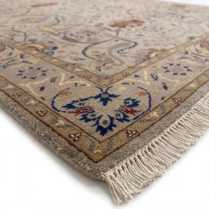 May include: Close-up of a beige area rug with an intricate floral pattern. The rug features a detailed border with blue, red, and green accents. Cream-colored fringe adds a decorative touch to the edge of the rug.