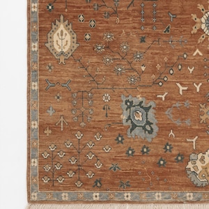 May include: A rectangular area rug featuring a warm terracotta brown background with a repeating pattern of stylised floral and geometric motifs in shades of slate blue, cream, and charcoal. The rug has a decorative border and white fringe.