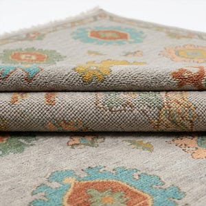 May include: Close-up of a rolled-up, light gray rug with a floral pattern. The rug features a repeating design of teal, orange, yellow, and green floral motifs. The edges of the rug are visible, showing the texture and thickness of the material.