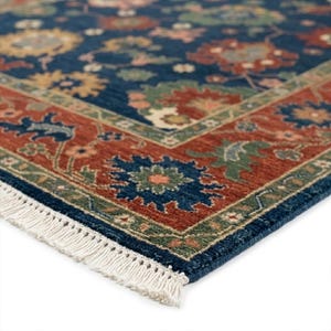 May include: Close-up of a blue and rust-colored area rug with a detailed floral pattern. The rug features a dark blue field with a repeating floral design in shades of red, green, and yellow. A rust-colored border frames the design, and the rug has white fringe.