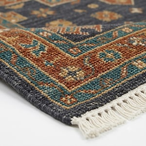 May include: A rectangular area rug featuring a dark charcoal grey base with a detailed border in shades of rust orange, teal, and cream. The rug has a textured, woven surface and is finished with short, off-white fringe along the edge.