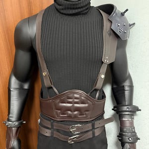 Puede incluir: Una camiseta negra de cuello alto con un arnés, cinturón y puños de cuero marrón. La armadura del hombro es gris con púas. El disfraz se exhibe en un maniquí.