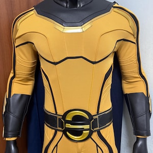 May include: A yellow and black superhero costume on a mannequin. The costume has a yellow bodysuit with black detailing, a black belt with a gold emblem, and black gloves. A dark blue cape is visible at the back.