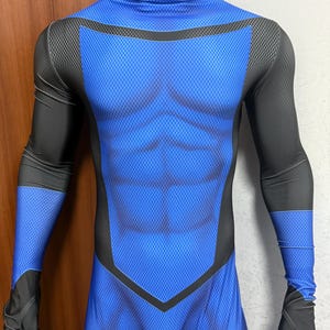 Puede incluir: Un disfraz de superhéroe azul y negro exhibido en un maniquí. El disfraz presenta un torso azul con un patrón texturizado y detalles negros en los hombros, brazos y guantes. El diseño incluye una apariencia de abdominales simulados.