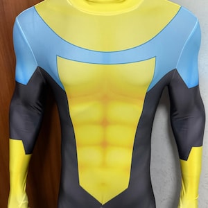 Può includere: Un costume da supereroe giallo, blu e nero con collo alto. Il costume presenta un busto giallo con un design blu sul petto e sulle spalle, maniche nere e guanti gialli. Il costume ha una cerniera frontale.