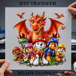 May include: A DTF transfer sheet featuring a large orange dragon with wings spread, positioned behind a group of five cartoon puppies in colorful uniforms. The text DTF TRANSFER and READY TO PRESS is printed at the top and bottom of the white sheet.