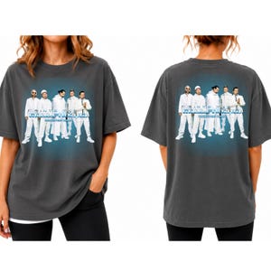 Puede incluir: Una camiseta de manga corta gris carbón con un estampado gráfico de los Backstreet Boys vestidos de blanco. El diseño incluye el texto "INTO THE MILLENNIUM" y "BACKSTREET BOYS" en una fuente con tonos azules. La camiseta se muestra por delante y por detrás.