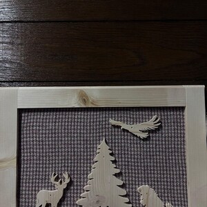 Może przedstawiać: Drewniana oprawiona grafika z wycięciami jelenia, sosny, orła i psa. Tło to wzorzysta tkanina w odcieniach brązu i szarości. Rama wykonana jest z jasnego drewna.