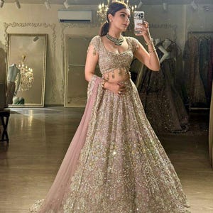 May include: A light pink and gold embellished lehenga, a traditional South Asian garment, with intricate embroidery and a matching dupatta. The outfit features a cropped top and a full, flowing skirt, perfect for formal events.