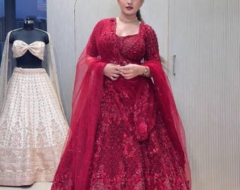 May include: A woman in a vibrant red, heavily embellished formal gown. The dress features intricate floral patterns and a matching sheer shawl. A mannequin displays a cream-colored, embroidered lehenga in the background. The setting appears to be a boutique or showroom.