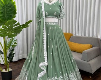 Può includere: Abito formale verde salvia con ricami bianchi intricati e una dupatta abbinata. L'abito presenta un top corto con maniche corte e una gonna fluida. Una collana di perline adorna il collo del manichino. L'abito è esposto su un manichino su sfondo neutro.