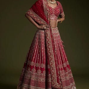 Op de afbeelding: Een levendig rode lehenga-ensemble met ingewikkelde gouden borduursels. De outfit omvat een bijpassende top, rok en een gedrapeerde sjaal. De rok is vloerlang en de top heeft korte mouwen. Het ontwerp is sierlijk en gedetailleerd.