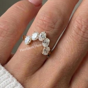 Puede incluir: Un anillo curvo con múltiples piedras preciosas transparentes. El anillo está en una mano que lleva un suéter blanco. Las piedras preciosas son ovaladas y en forma de pera, y el anillo tiene una banda dorada. Las palabras "Heirloom Hearts" están en las esquinas superior izquierda y derecha.