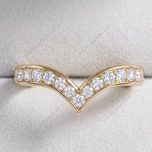 Puede incluir: Un anillo dorado en forma de V con una fila de piedras preciosas redondas y transparentes. El diseño del anillo se destaca sobre un fondo gris claro y suave, resaltando el brillo de las piedras.