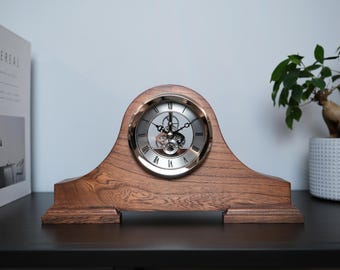 Può includere: Un orologio da mensola in legno con quadrante color argento e numeri romani. L'orologio ha un meccanismo a ingranaggi visibile ed è incastonato in una cornice di legno marrone ad arco. L'orologio è su una superficie scura.