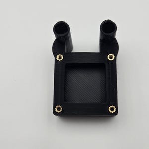 Peut inclure: Un composant noir imprimé en 3D avec deux protubérances cylindriques et une base rectangulaire. La base comporte quatre fixations dorées. L'objet est posé sur un fond blanc.