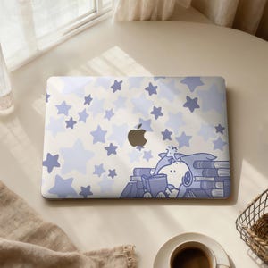 Puede incluir: Una funda para portátil con un fondo beige claro y estrellas azules de diferentes tamaños. El diseño incluye una ilustración de dibujos animados de un perro leyendo libros. El logotipo de Apple es visible.