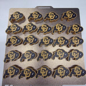 May include: A collection of black and gold buffalo-shaped objects, each featuring the CU logo. The objects are arranged in rows on a gold-colored surface. The buffalo shapes have a textured appearance.