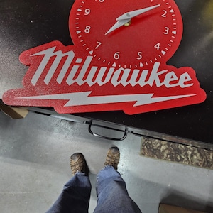 May include: A red wall clock with white hands and a white face. The clock is in the shape of the Milwaukee logo, with the brand name and a lightning bolt design. The clock is on a black surface.