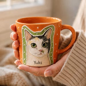 Peut inclure: Un mug en céramique orange orné d'un portrait peint à la main d'un chat calico aux yeux verts. Le chat est encadré par une bordure vert clair à pois blancs, et le nom Yuki est inscrit en lettres noires sous le menton du chat sur la tasse.