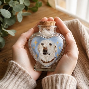 May include: A heart-shaped glass bottle with a cork stopper containing a hand-painted portrait of a cream-colored dog. The dog has a brown collar and is set against a light blue background with yellow four-pointed star accents painted on the glass.