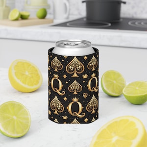 Puede incluir: Un enfriador de latas negro con un diseño dorado de picas, coronas y "Q". El diseño se repite en todo el enfriador. Una lata plateada está dentro. Limones y limas en rodajas están en primer plano.