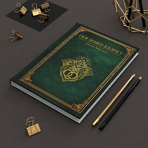 May include: A green journal with gold accents and the text "OUR SHARED JOURNEY Lifestyle Reflections." The cover features a gold art deco design with interlocking rings. Gold pencils and binder clips are also visible.