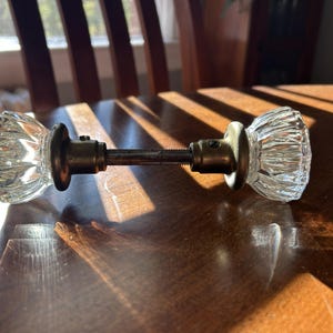 May include: A door knob set featuring two clear, faceted glass knobs with a fluted design. The knobs are connected by a central metal spindle and include two circular, dark bronze-toned metal rosettes with visible set screws.