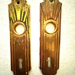 Może przedstawiać: Dwie zabytkowe mosiężne płytki do klamek. Każda płytka ma prostokątny kształt z okrągłym otworem i otworem na klucz. Górna część każdej płytki ma wzór w kształcie wachlarza. Mosiądz ma zużyty wygląd.