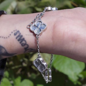 Puede incluir: Una pulsera de plata con tres colgantes en forma de trébol y un anillo. Los colgantes tienen un diseño texturizado y están rodeados de pequeños detalles brillantes. La pulsera se muestra en una muñeca.