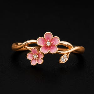 以下が含まれることがあります： ピンクのエナメルで仕上げた2輪の桜の花と小さな葉のモチーフがあしらわれたゴールドトーンのリング。それぞれの花の中央には小さな透明な石がセットされ、葉の部分には3つの小さな透明な石が施されており、細くカーブしたリングバンドが特徴です。