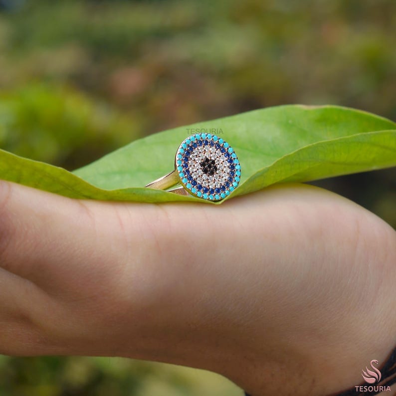May include: Gold-toned ring featuring a circular design with a central black dot, surrounded by white, blue, and turquoise stones. The ring is set against a green leaf, showcasing its vibrant colors and intricate details. The ring's design resembles an eye.