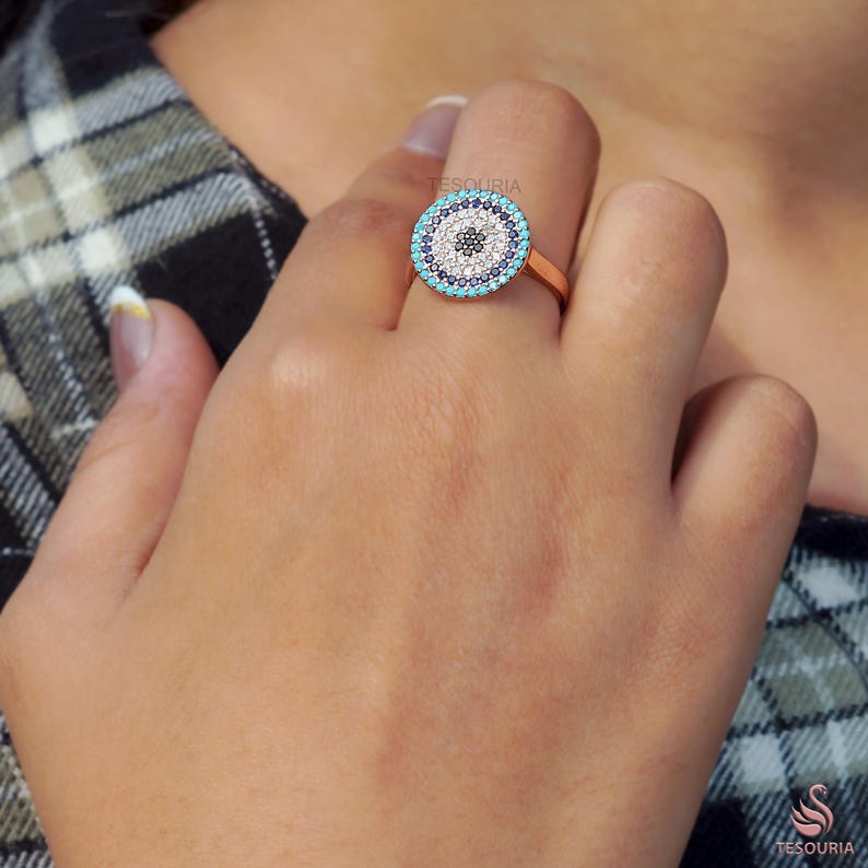 May include: A rose gold ring featuring a circular design with a central black dot surrounded by white, turquoise, and navy blue stones. The ring is on a person's finger, showcasing the intricate details of the jewelry.