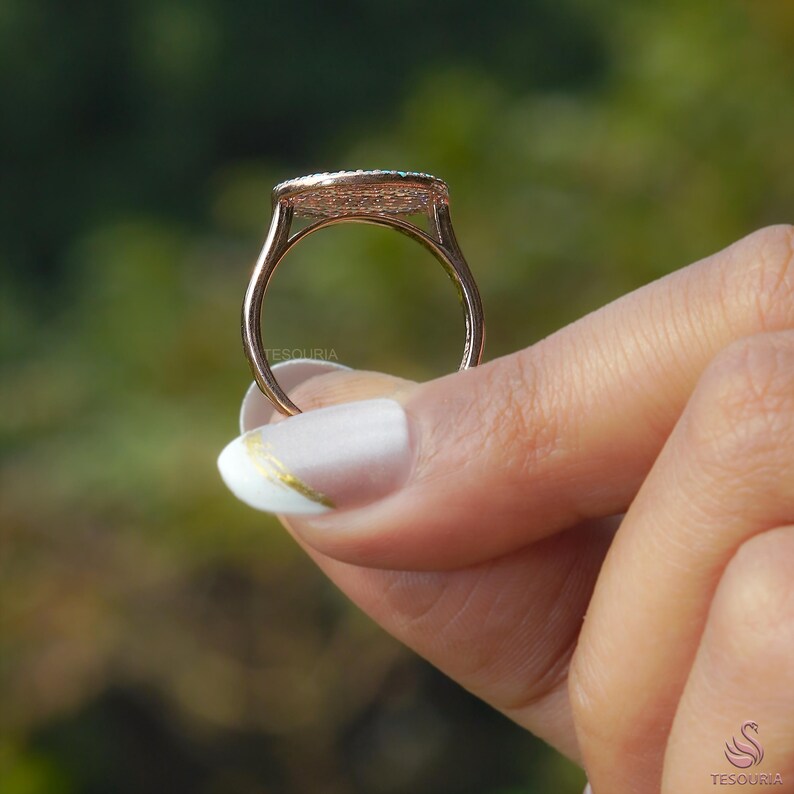 May include: A rose gold ring with a rectangular, emerald-cut gemstone setting. The ring is held by a hand with a white manicure and gold accent. The ring's band is thin and delicate, and the setting is detailed.