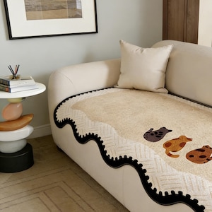 May include: A cream-colored sofa with a patterned cushion featuring three cartoon animal figures in brown and tan. The sofa has a black wavy trim and a matching cream-colored pillow. A small side table with stacked decorative stones is visible.
