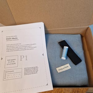 Può includere: Una scatola di cartone aperta contenente materiali da cucito. All'interno, un pezzo di tessuto blu piegato, un elastico nero, una bobina di filo blu e un'etichetta con la scritta "Sewing with Emilia". È visibile anche un foglio modello.