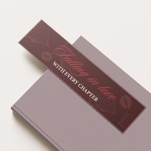 Könnte beinhalten: Ein weinrotes Lesezeichen mit dem Text "Falling in love with every chapter" in einer Schreibschrift, das auf einem geschlossenen Buch liegt. Das Lesezeichen zeigt Buch- und Lippenillustrationen.