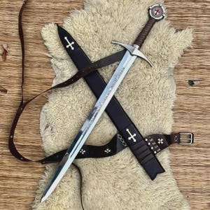 May include: A medieval sword with a silver blade and a brown leather handle, on a white fur rug. The sword is in a black sheath with a brown leather belt. The handle has a red and white emblem.