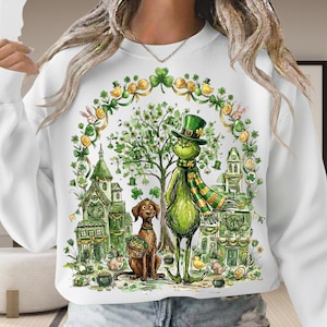 May include: White sweatshirt featuring a whimsical St. Patrick's Day design. The graphic includes a green character with a hat and scarf, a brown dog, and green buildings. The design is surrounded by shamrocks and gold coins.
