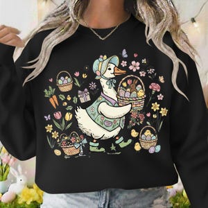 May include: Black sweatshirt featuring a cartoon goose dressed for Easter, carrying a basket of eggs and a bunny. The design includes flowers, butterflies, and additional baskets with eggs and chicks. The goose wears a bonnet and vest.