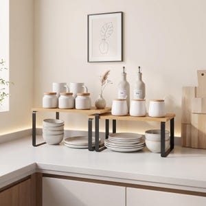 May include: Two tiered kitchen counter shelves with black metal frames and light wood surfaces. The shelves hold white ceramic canisters with wooden lids, white bowls, white plates, and two glass oil bottles with silver pour spouts.