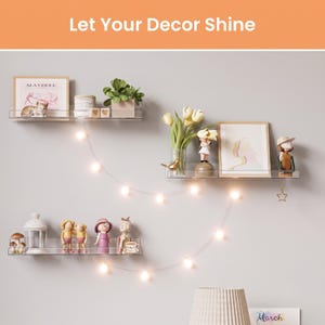 May include: Three clear acrylic floating wall shelves arranged in a staggered pattern. The shelves hold various decorative items including framed art, small figurines, a potted plant, a white lantern, and a string of warm white LED fairy lights.