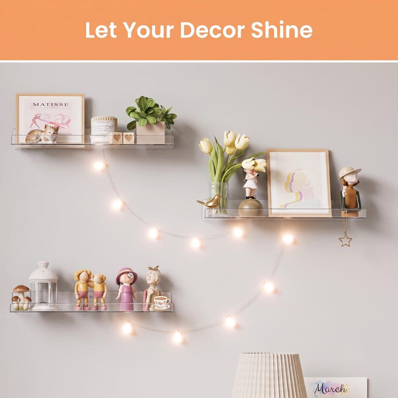 May include: Three clear acrylic floating wall shelves arranged in a staggered pattern. The shelves hold various decorative items including framed art, small figurines, a potted plant, a white lantern, and a string of warm white LED fairy lights.