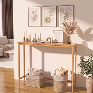 May include: A tall, rectangular bamboo console table with four slender legs. On the table are three taper candles in holders, an open book, a beige ceramic vase, a framed print, and a glass vase filled with dried pampas grass plumes.